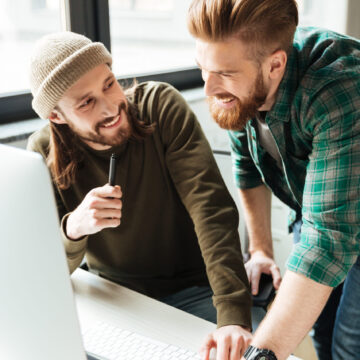 Photo of young happy men colleagues in office using computer. Looking aside.